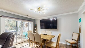A dining area with a wooden table and chairs, a television mounted on the wall, and a sliding glass door leading to an outdoor space.
