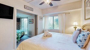 Un dormitorio con cama, ventilador de techo, ventana con cortinas, televisor montado en la pared y puerta que da a otra habitación.