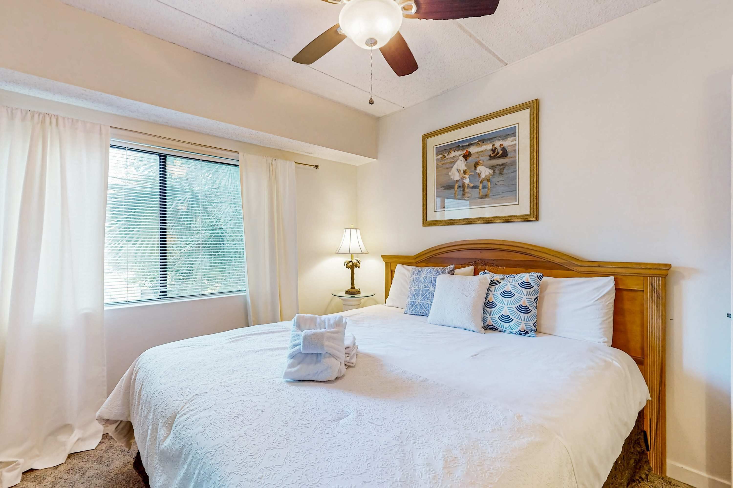 A hotel room with a bed, a ceiling fan, a window with curtains, a bedside lamp, and a framed picture on the wall.