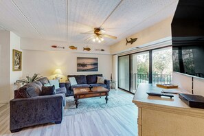 A living room with a sofa set, a coffee table, a ceiling fan, and a large window with a view. - Close to beach with pool, tennis, pool  & private balcony (Hilton Head)