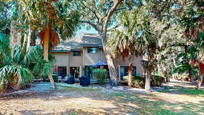 Una casa a due piani con un portico coperto, circondata da alberi e terreno sabbioso.