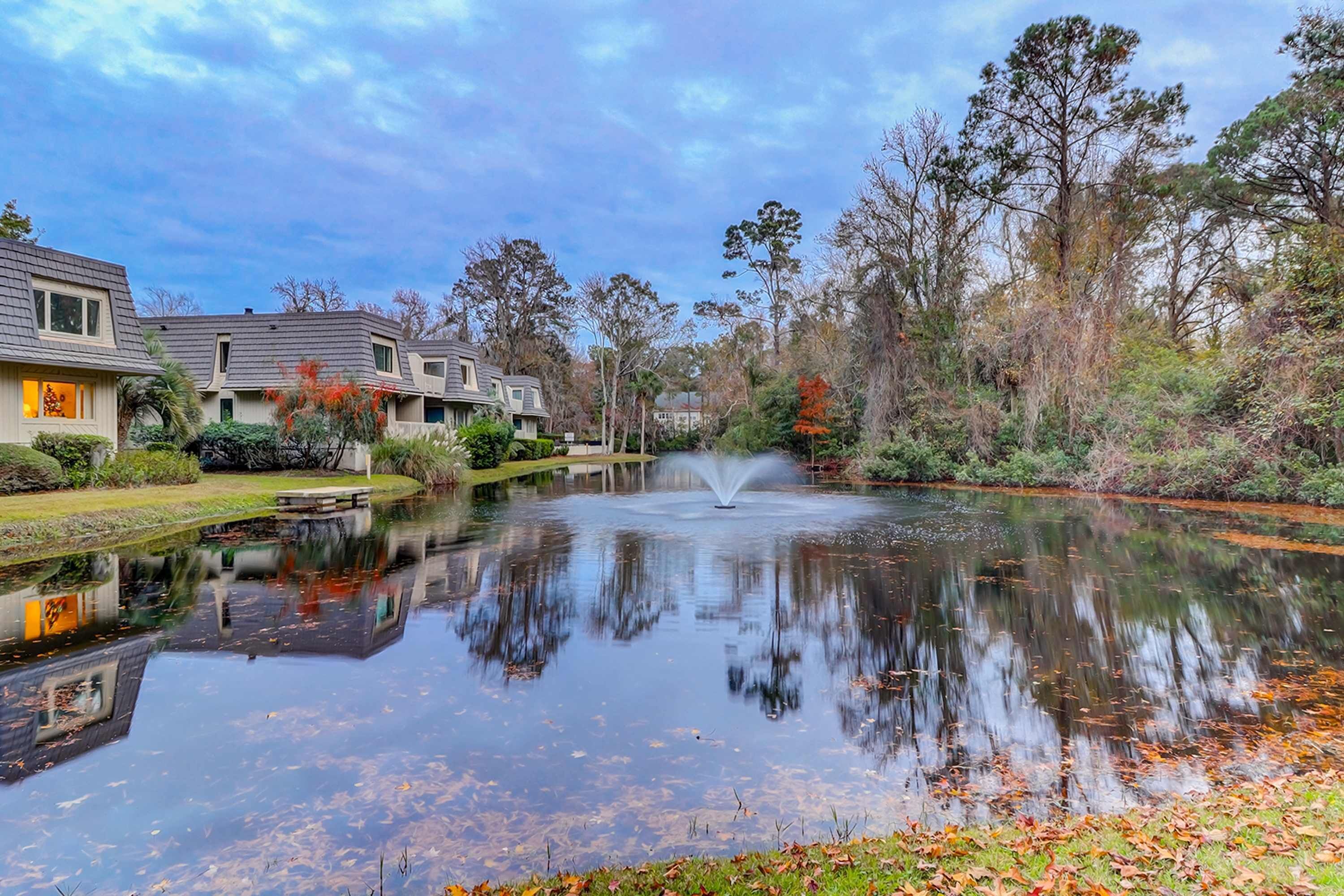 A tranquil pond with a fountain, surrounded by trees and residential buildings.