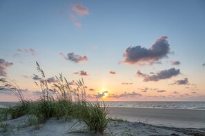 Una playa con dunas de arena y pastos en primer plano, y un atardecer sobre el océano.