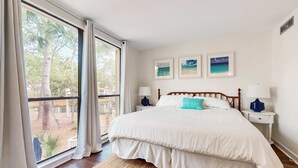 A bedroom with a large window, a bed with white bedding, two bedside tables with lamps, and three framed pictures on the wall. - Coastal condo with private grill and shared pool, nearby beach access (Hilton Head)