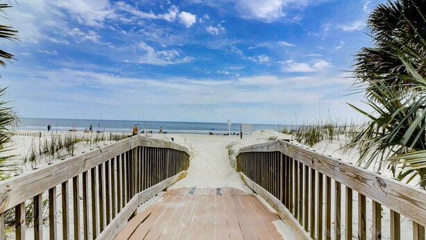 Ein hölzerner Steg führt zu einem Strand unter blauem Himmel.