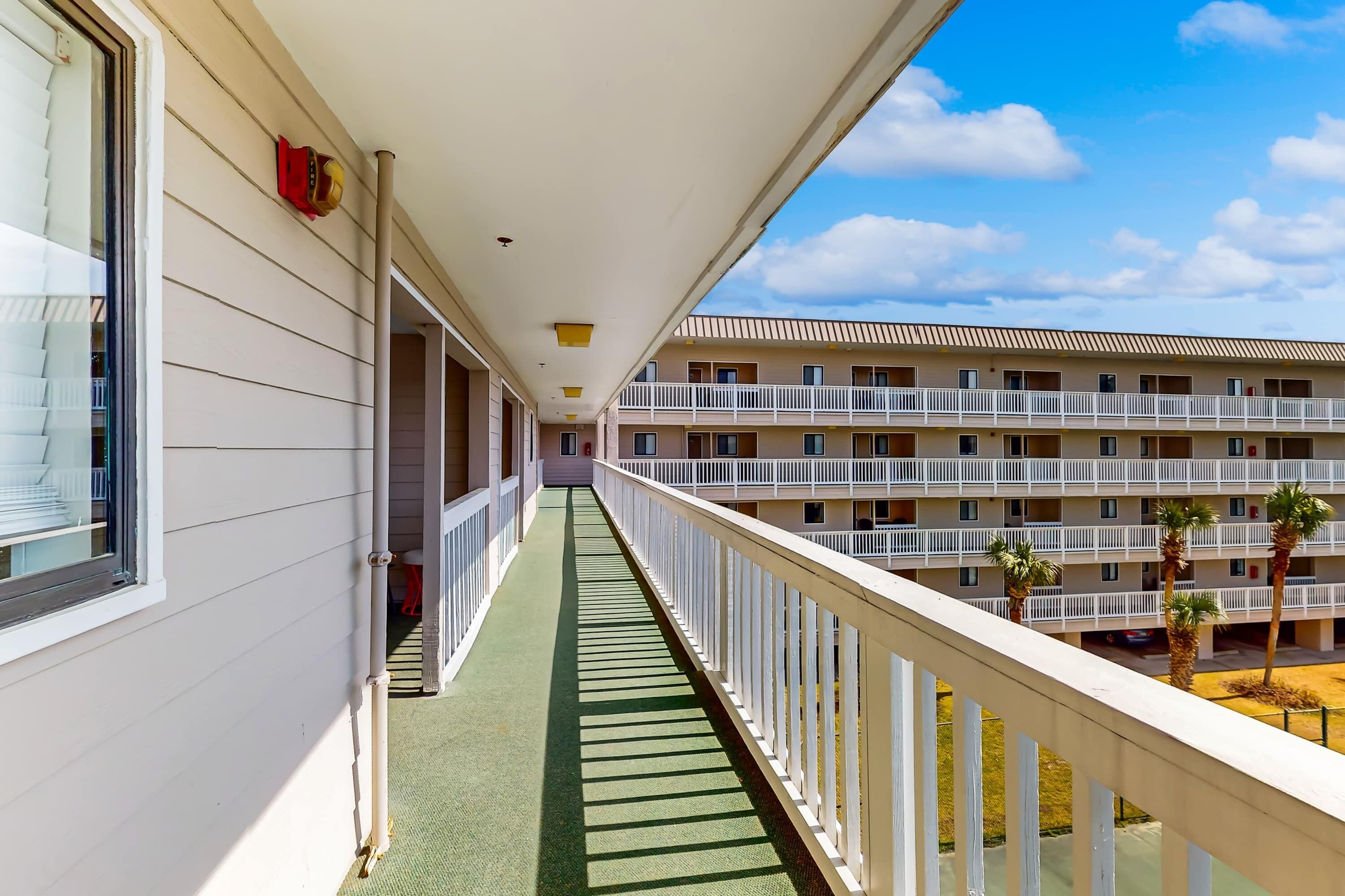 A balcony with a green mat, white railings, and a view of a multi-story building with balconies and palm trees.