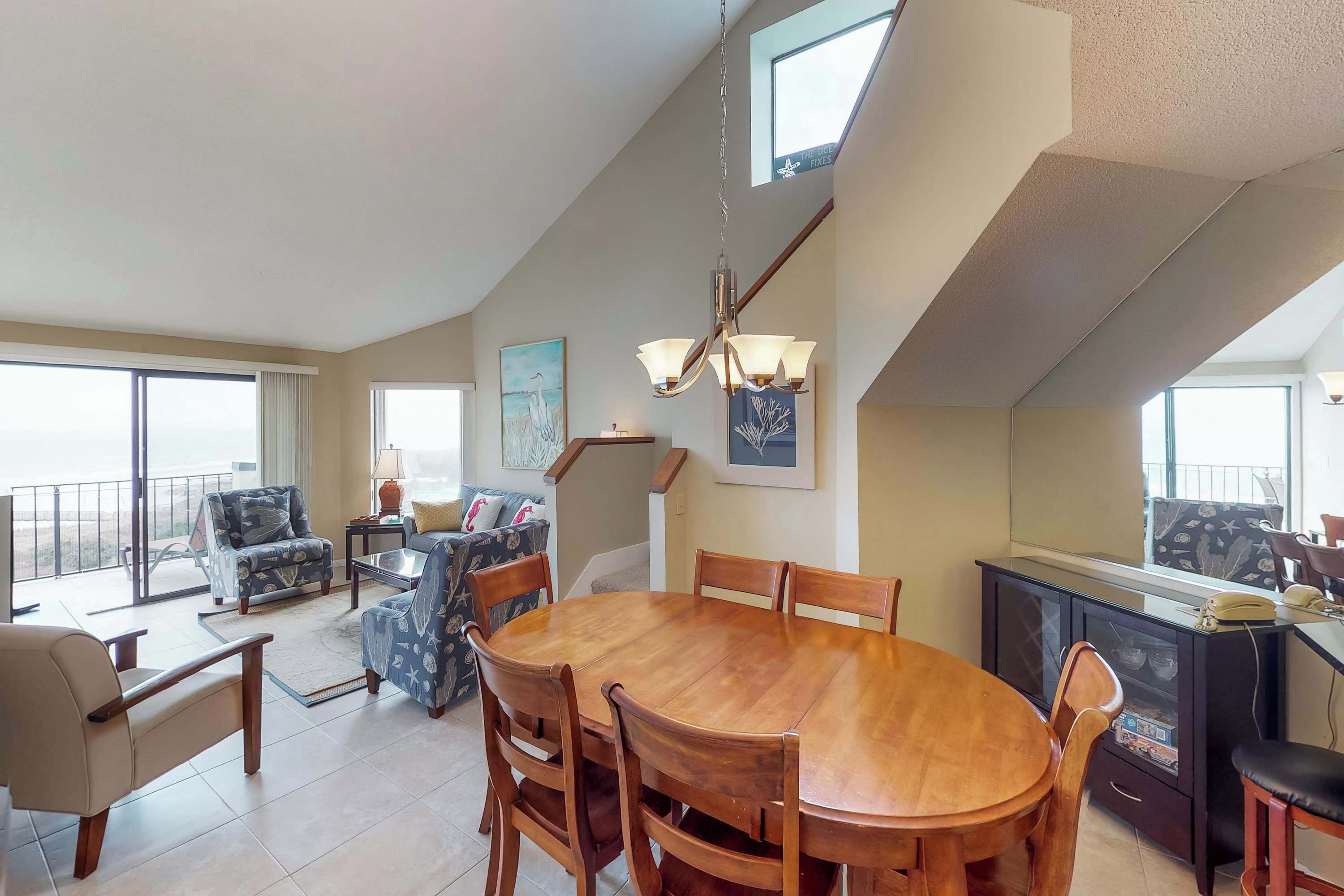 A spacious living area with a large wooden dining table, comfortable seating, a balcony with a view, and a modern kitchen with a refrigerator.
