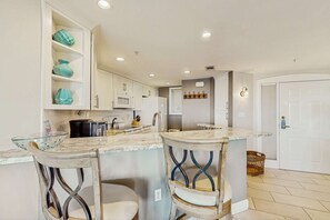 A kitchen with white cabinets, a microwave, a refrigerator, a sink, and a bar area with two stools. - A spacious condo with community pool, hot tub, and oceanfront views (Hilton Head)