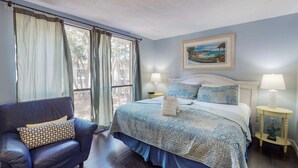 Un dormitorio con cama, una silla, una mesita de noche, una lámpara y un cuadro en la pared.