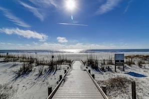 Ein Holzsteg führt zu einem Strand bei strahlend blauem Himmel, mit einem Schild, das einen Bereich für Strandordnungen kennzeichnet.