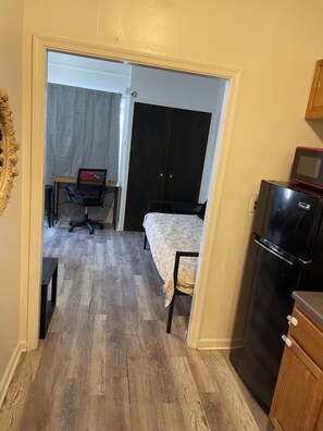 Ein kleines Zimmer mit einem Bett, einem Schreibtisch, einem Stuhl, einem Kühlschrank und einer Tür zu einem weiteren Raum.