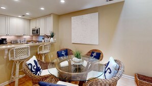 Uma área de refeições com uma mesa de vidro rodeada por cadeiras de vime, uma cozinha ao fundo e um quadro branco na parede.