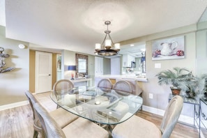 A dining area with a glass table, wicker chairs, a chandelier, and a kitchen in the background. - Spacious villa with pool, hot tub, playground & cute decor - steps from beach (Hilton Head)