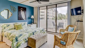 A bedroom with a bed, a ceiling fan, a large window, a TV, and a balcony with outdoor furniture. - Resort Condo with Al Fresco Dining, Shared Pool, Hot Tub & Beach Access (Hilton Head)