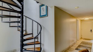 A spiral staircase with wooden steps and black metal railings, a framed picture on the wall, and a door at the end of a hallway with a doormat. - Resort Condo with Al Fresco Dining, Shared Pool, Hot Tub & Beach Access (Hilton Head)