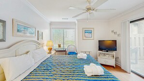 Ein Hotelzimmer mit Bett, Deckenventilator, Fernseher, Schreibtisch mit Lampe und Blick nach draußen.