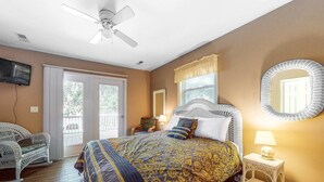 Ein Schlafzimmer mit einem Bett, einem Deckenventilator, einem Fernseher, einem Fenster mit Vorhängen, einem Korbstuhl und einem kleinen Tisch mit einer Lampe.