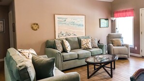 Ein Wohnzimmer mit einem grünen Sofa, einem Glastisch, einem beigen Sessel und einem Bild von einer Strandansicht.