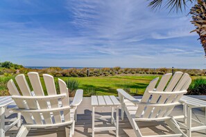 A deck with Adirondack chairs and a table, overlooking a grassy field and the ocean. - First Floor, Oceanfront Condo with Free WiFi, Balcony, Shared Hot Tub, Pool, Sauna (Hilton Head)