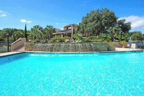 A swimming pool with a stone waterfall feature, surrounded by lush greenery and a building with a curved roof. - Ocean-View Condo with Beach Access, Tennis, Playground, Shared Pool & Hot Tub (Hilton Head)