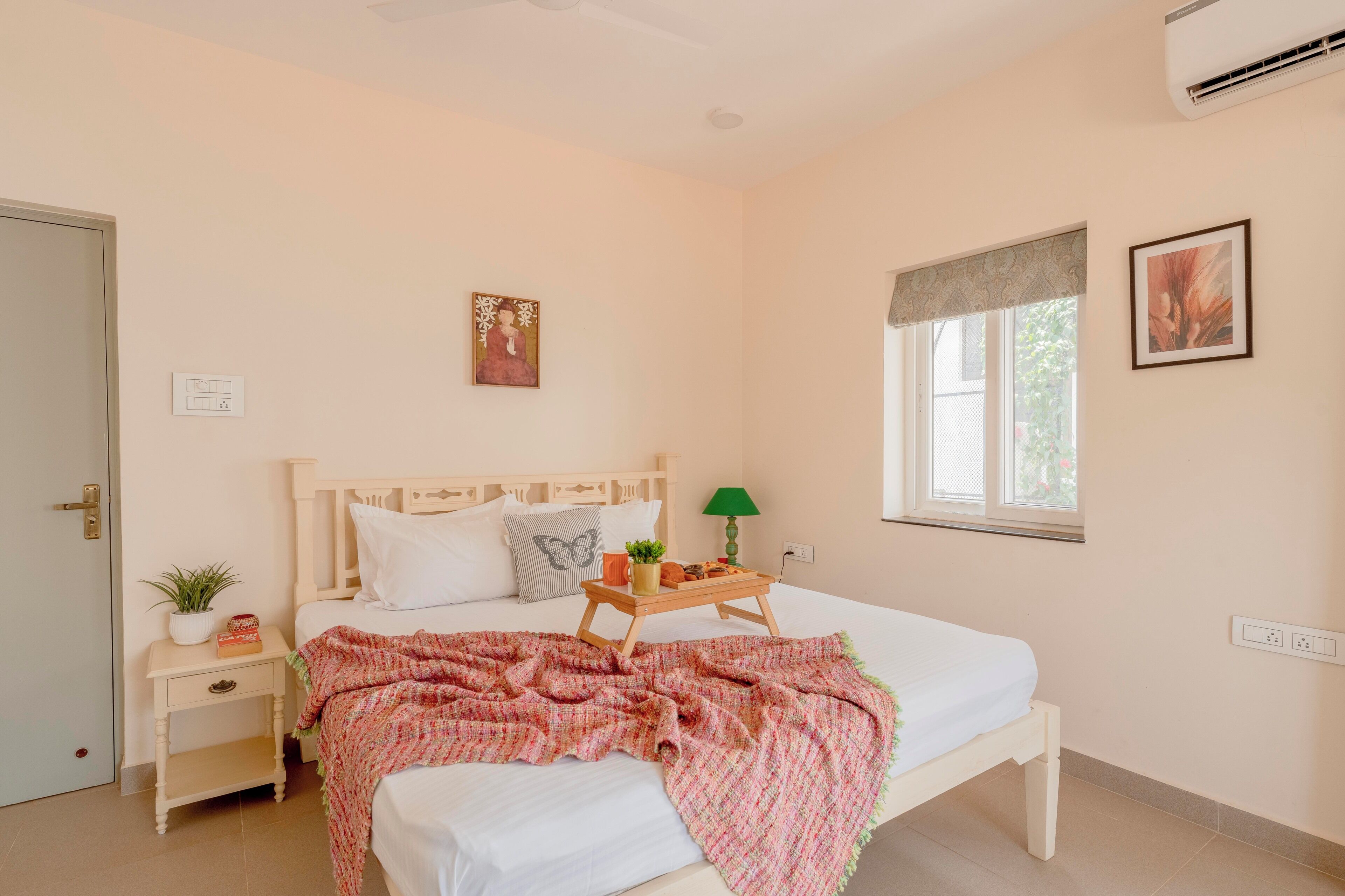 A bedroom with a bed, bedside table, lamp, window, and artwork.