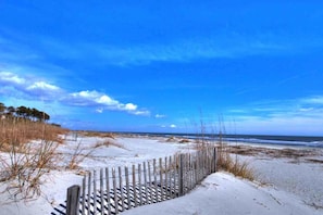 Ein sandiger Strand mit einer Holzlattenzaun, blauem Himmel und dem Ozean im Hintergrund.