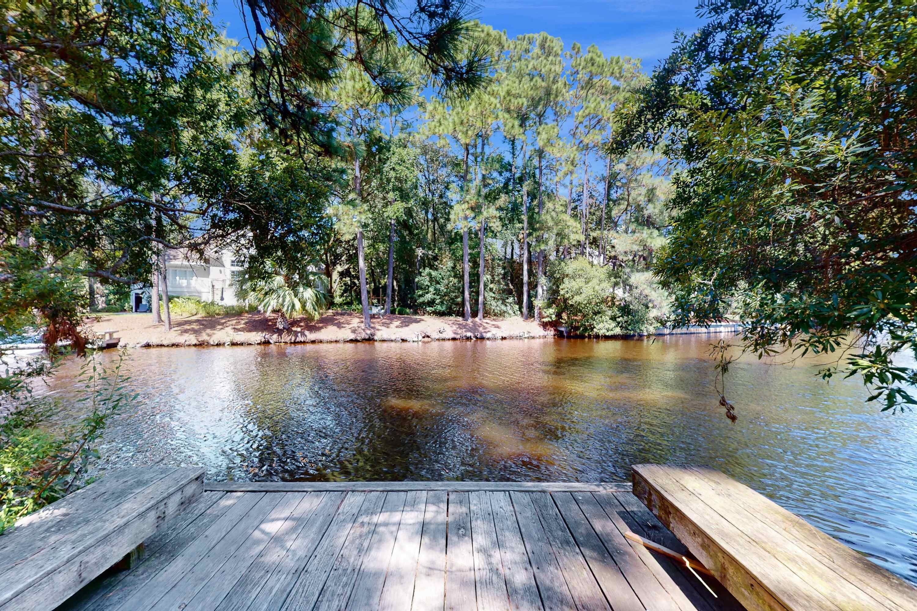 Une jetée en bois qui s’avance sur un lac paisible, entouré d’arbres, avec une maison en arrière-plan.
