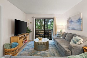 Ein Wohnzimmer mit Fernseher, einer Couch, einem Couchtisch und Blick durch eine Glastür auf einen Außenbereich.