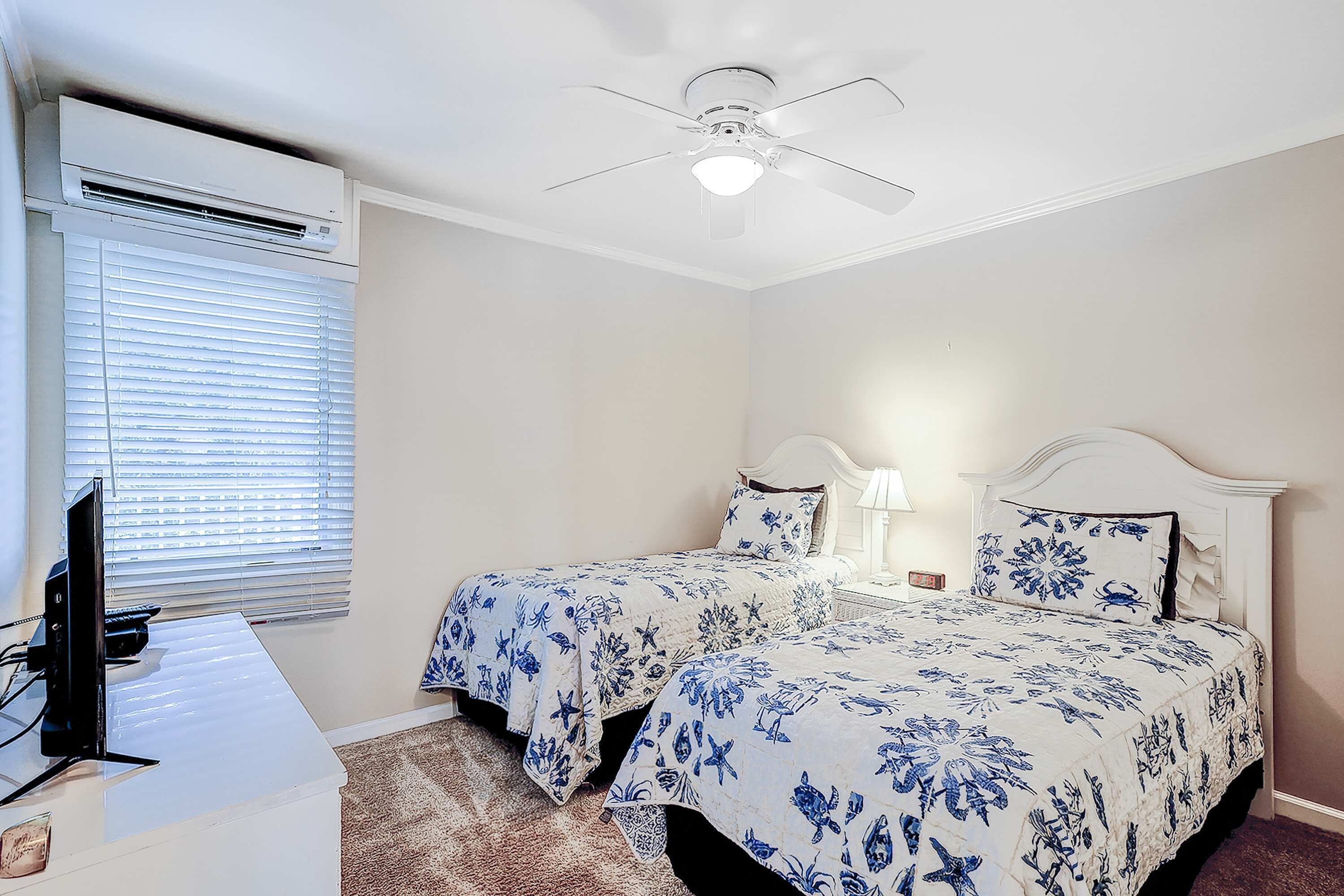 A bedroom with two beds, an air conditioning unit, a ceiling fan, a desk with a computer, and a window with blinds.
