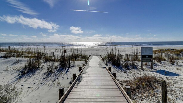Ein Holzsteg führt zu einem Strand bei blauem Himmel und einem Schild.