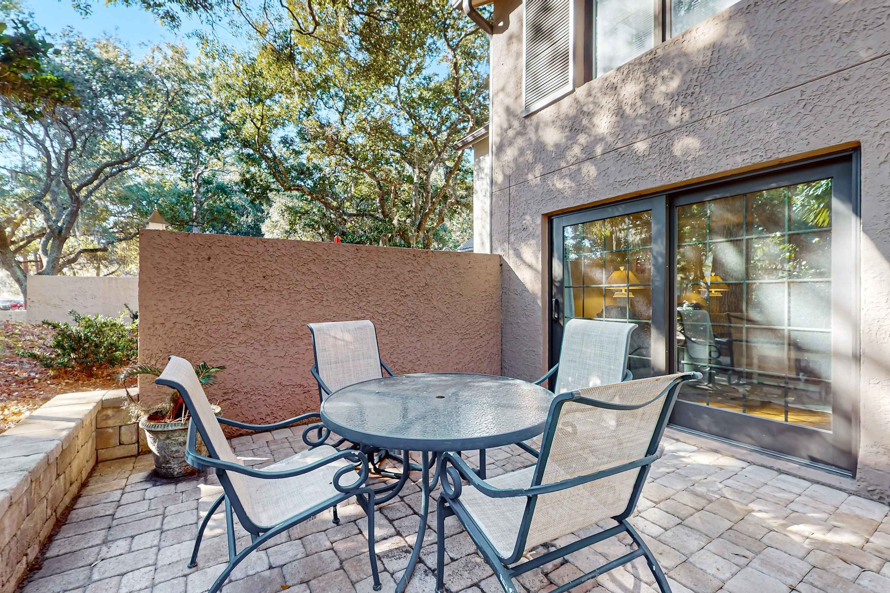 Un patio exterior con una mesa redonda y cuatro sillas, una planta en maceta, y un edificio con pared texturizada y ventanas amplias.