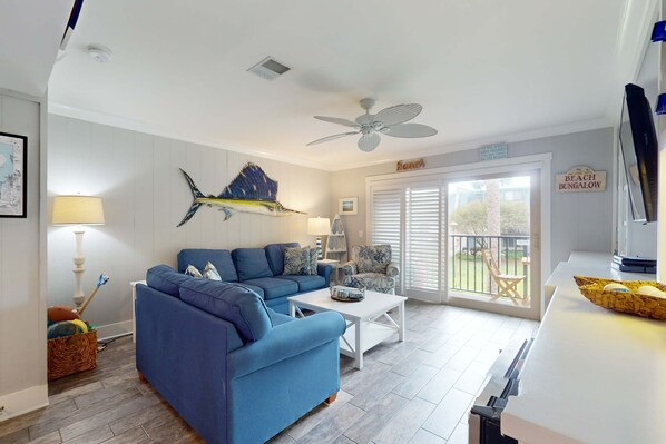 Ein Wohnzimmer mit blauem Sofa, weißem Couchtisch, Deckenventilator und Blick ins Freie.