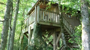 A wooden treehouse with a balcony and stairs, surrounded by forest.