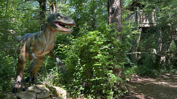 A dinosaur statue in a forested area with trees and a stone structure.