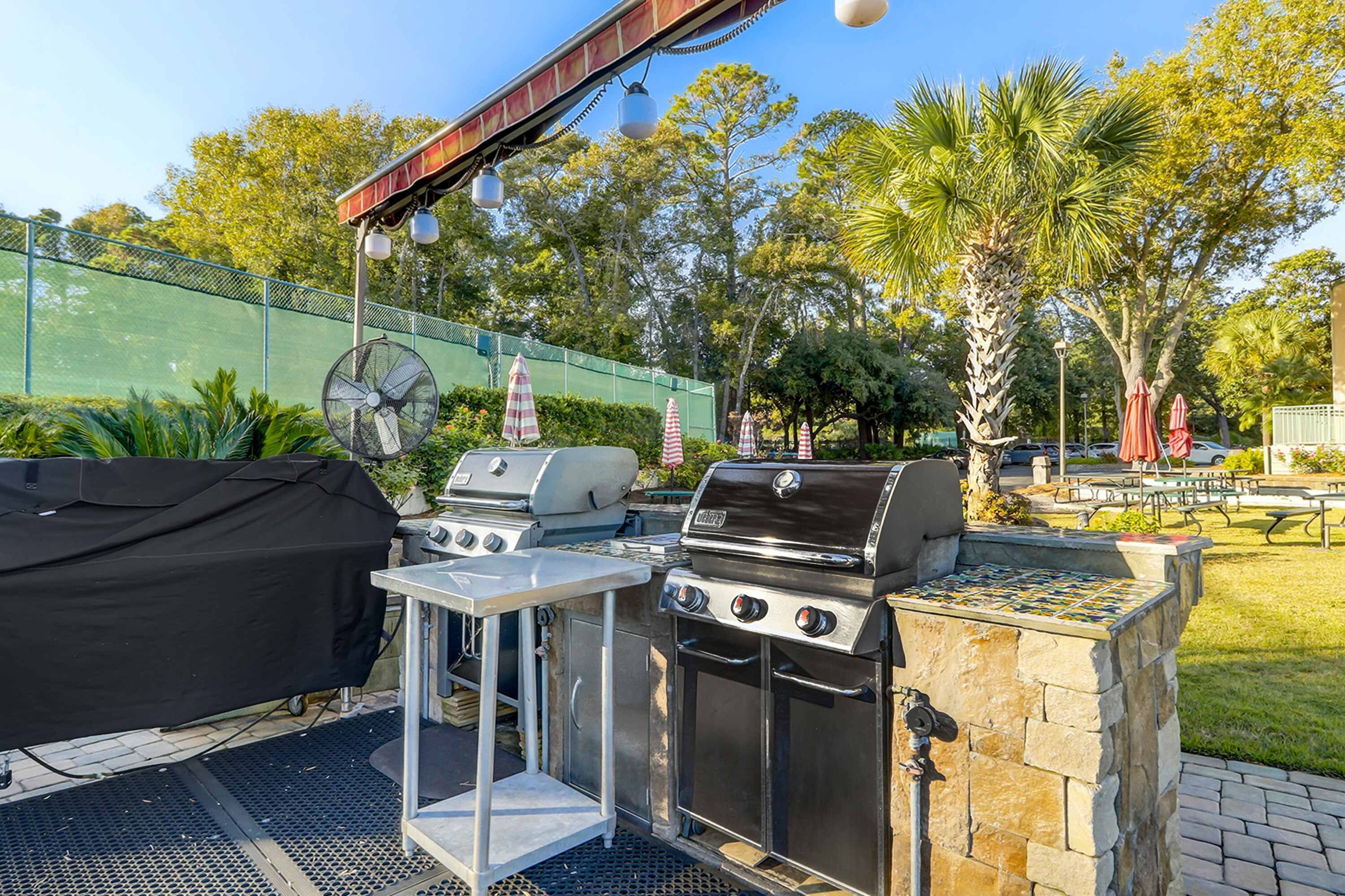 Una zona de parrilla al aire libre con una parrilla cubierta, un ventilador y una mesa con funda para parrilla.