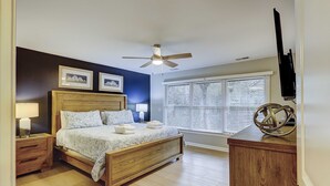 A bedroom with a wooden bed, bedside tables, a ceiling fan, a window with blinds, and a TV mounted on the wall. - Breezy resort home located in Sea Pines Resort near marina & restaurants (Hilton Head)
