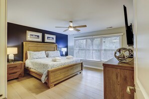 Ein Schlafzimmer mit einem hölzernen Bett, Nachttischen, einem Deckenventilator, einem Fenster mit Jalousien und einem an der Wand montierten Fernseher.