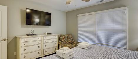Ein Schlafzimmer mit Bett, Deckenventilator, Fernseher, Kommode, Stuhl und Fenster mit Jalousien.