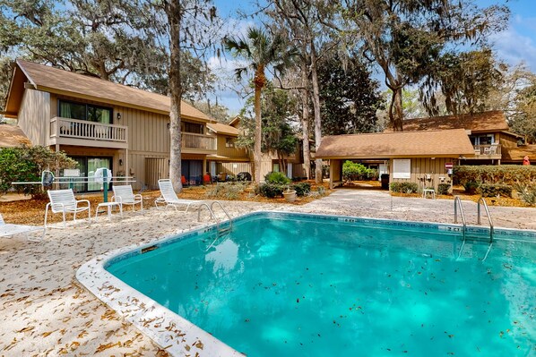 A pool area with lounge chairs, a wooden building with balconies, and surrounding trees. - Pool-view condo near beach with non-heated pool, golf, tennis, grill (Hilton Head)
