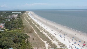 Vista aérea de uma praia com uma extensa faixa de areia, alguns edifícios à esquerda e um céu claro.