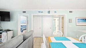 Quarto com televisão, sofá, mesa de jantar e vista para a praia através de uma porta.