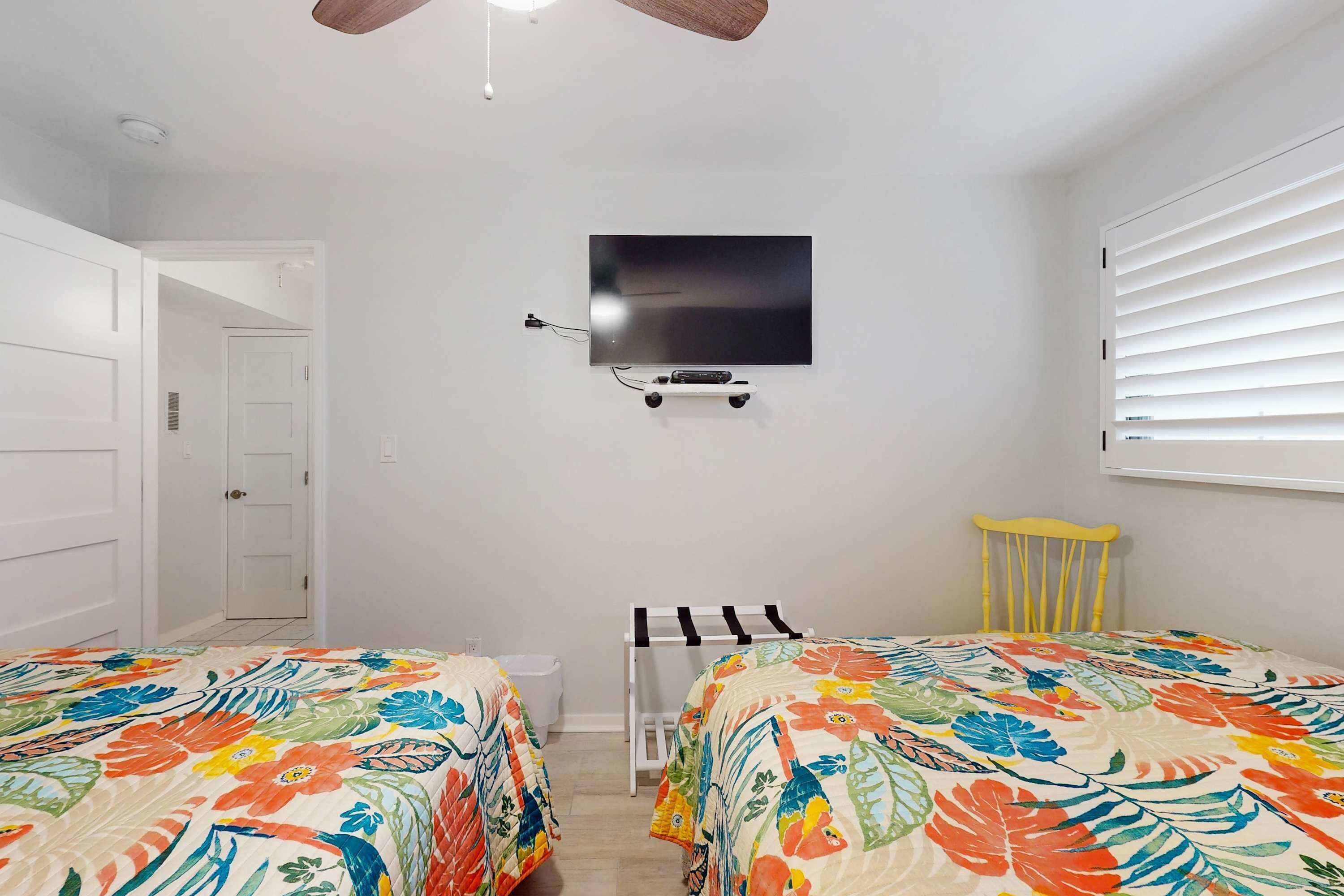 A bedroom with two beds, a ceiling fan, a TV mounted on the wall, and a window with blinds.