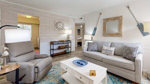 A living room with a sofa, a coffee table, a lamp, a bookshelf, and a wall clock. - Relaxing condo with heated chilled pool, easy beach access, patio, & W D (Hilton Head)