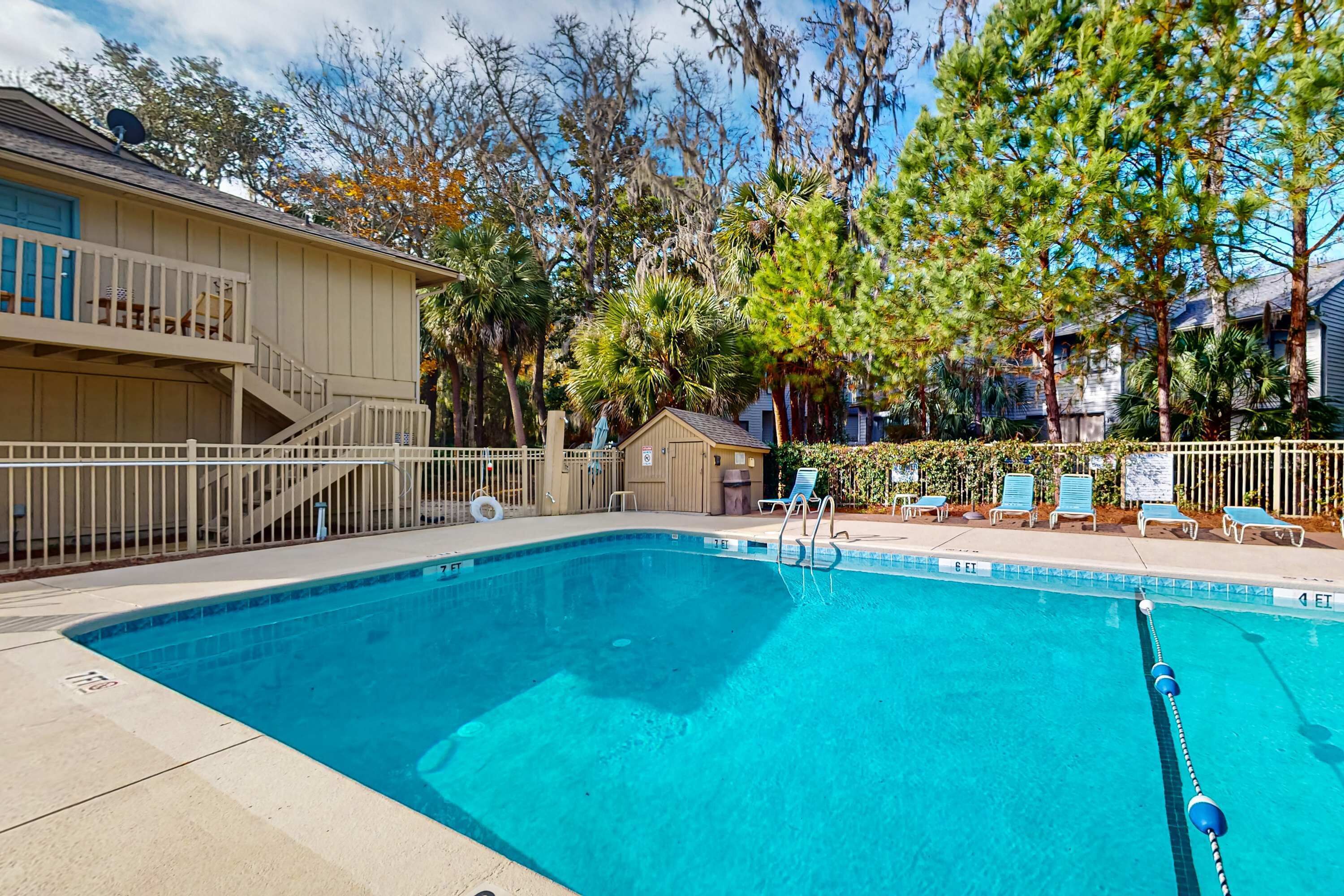 A swimming pool with a white fence, lounge chairs, and a building with a balcony.