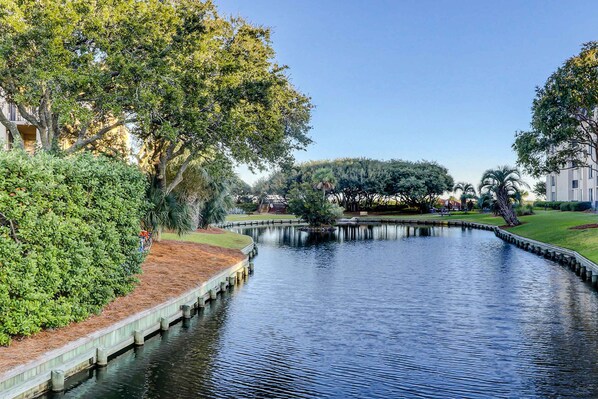 A calm water body with a wooden dock, surrounded by lush greenery and trees, with buildings in the background. - Tropical Condo with Pool, Hot Tub, Lagoon Views from Balcony - Beach Access (Hilton Head)