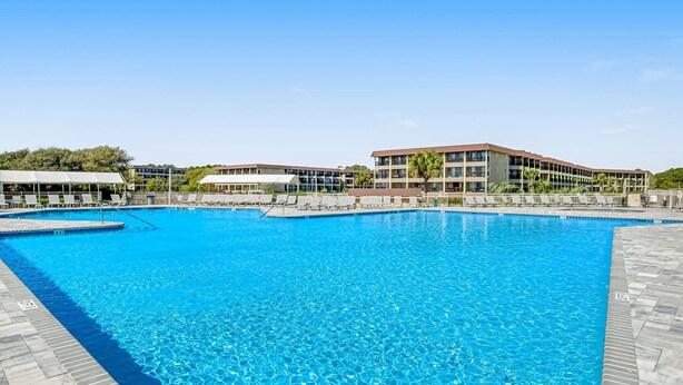 A large swimming pool with clear blue water, surrounded by a tiled deck and a building with multiple floors and balconies. - 1st floor condo near the beach with pool, tennis, balcony, gym, streaming, WD (Hilton Head)