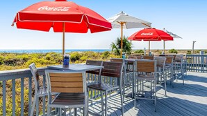 Outdoor seating with red Coca-Cola umbrellas, wooden tables, and metal chairs on a wooden deck with a view of the beach. - 1st floor condo near the beach with pool, tennis, balcony, gym, streaming, WD (Hilton Head)
