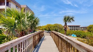 A wooden boardwalk leading to a building with balconies, surrounded by palm trees and greenery. - 1st floor condo near the beach with pool, tennis, balcony, gym, streaming, WD (Hilton Head)