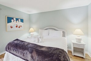 A bedroom with a bed, two bedside tables with lamps, a wall-mounted painting, and a window. - Delightful Condo with WiFi, Pool, Tennis - Beach Across the Street (Hilton Head)