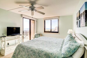 Une chambre avec un lit, un ventilateur de plafond, un téléviseur posé sur un meuble blanc, une fenêtre donnant sur un paysage agréable et un balcon.